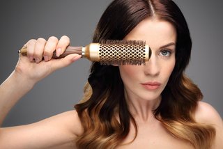 Portret kobiety z falowanymi ciemnymi włosami i jasnymi końcówkami. W dłoniach przed twarzą trzyma okrągłą szczotkę nadającą włosom objętości