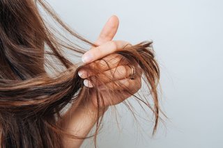 Zbliżenie na dłoń kobiety, która trzyma swoje włosy i pokazuje łamliwe końcówki. Włosy ma długie i brązowe