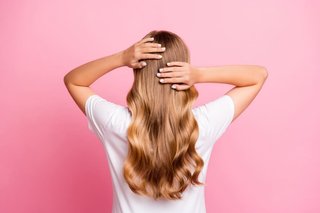 Odwrócona tyłem kobieta w białej koszulce, dotyka dłońmi falowanych, blond włosów